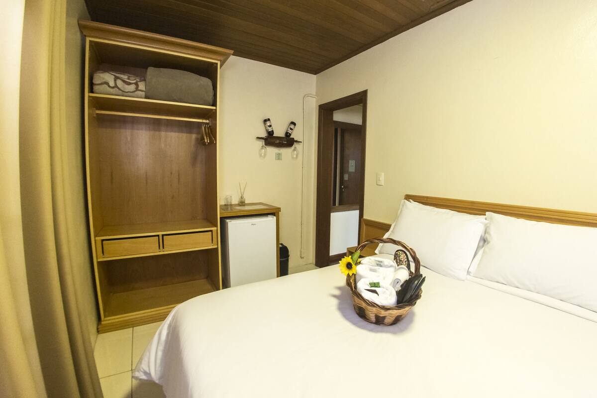 Ein Hotelzimmer mit einem Bett, einem Holzschrank, einem kleinen Schreibtisch mit Lampe und einem an der Wand befestigten Regal mit zwei Flaschen.