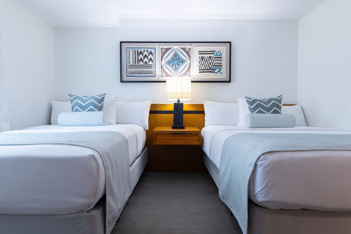 Ein Hotelzimmer mit zwei Betten, einem Nachttisch mit Lampe und einer Wand mit drei gerahmten Kunstwerken.