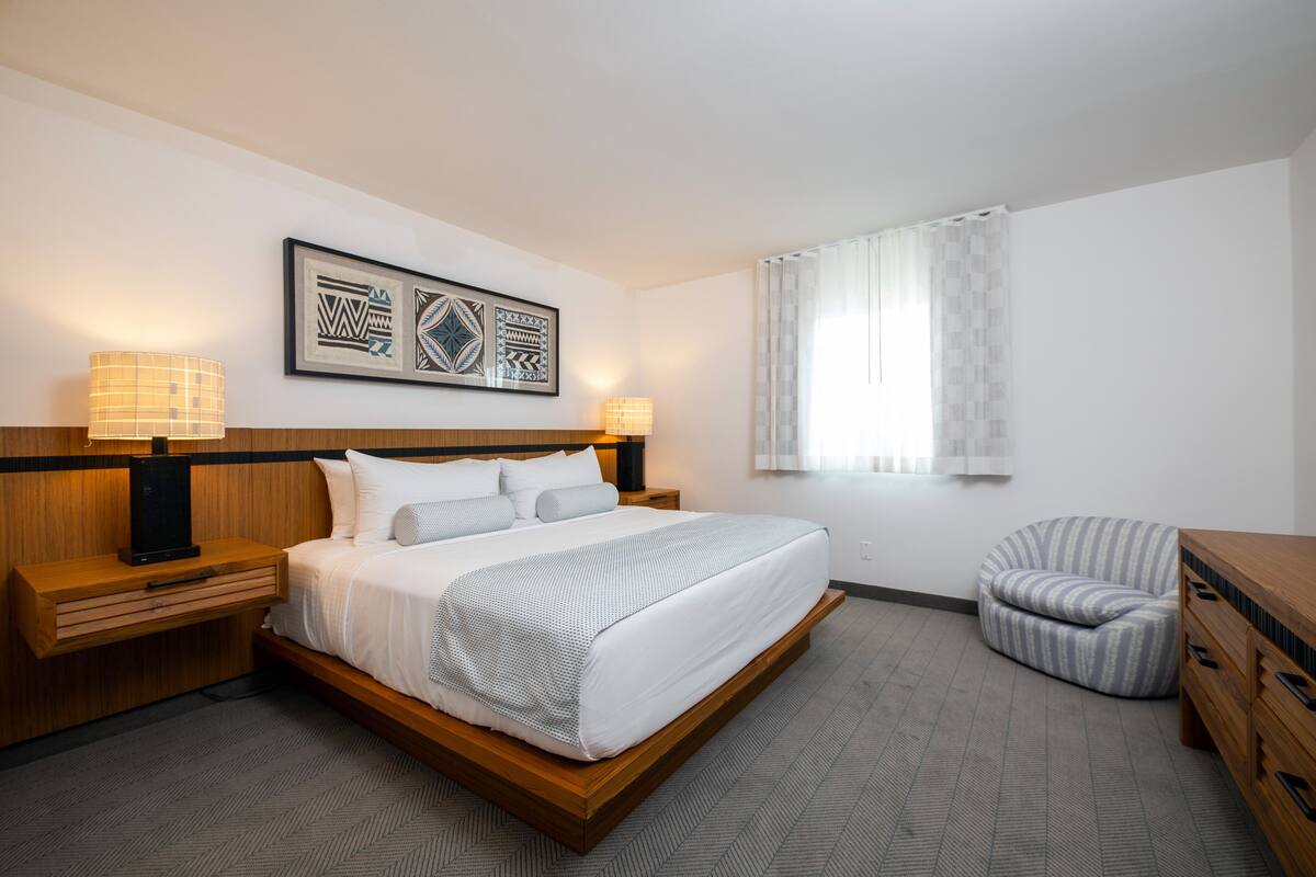 Ein modernes Hotelzimmer mit einem großen Bett, Nachttischlampen, einem Holznachttisch, einem Sessel und einem Fenster mit Vorhängen.