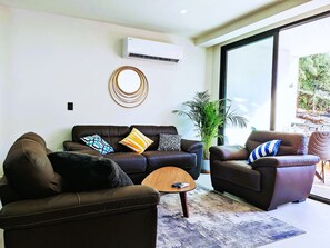 A modern living room with a brown leather sofa, a coffee table, and a large window providing a view of greenery outside.