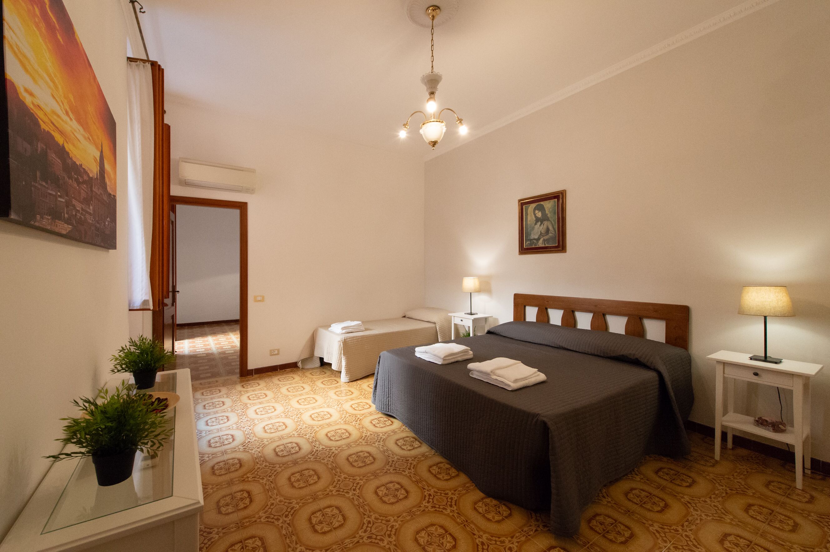 A hotel room with a bed, bedside tables, a nightstand, a potted plant, and a painting on the wall.