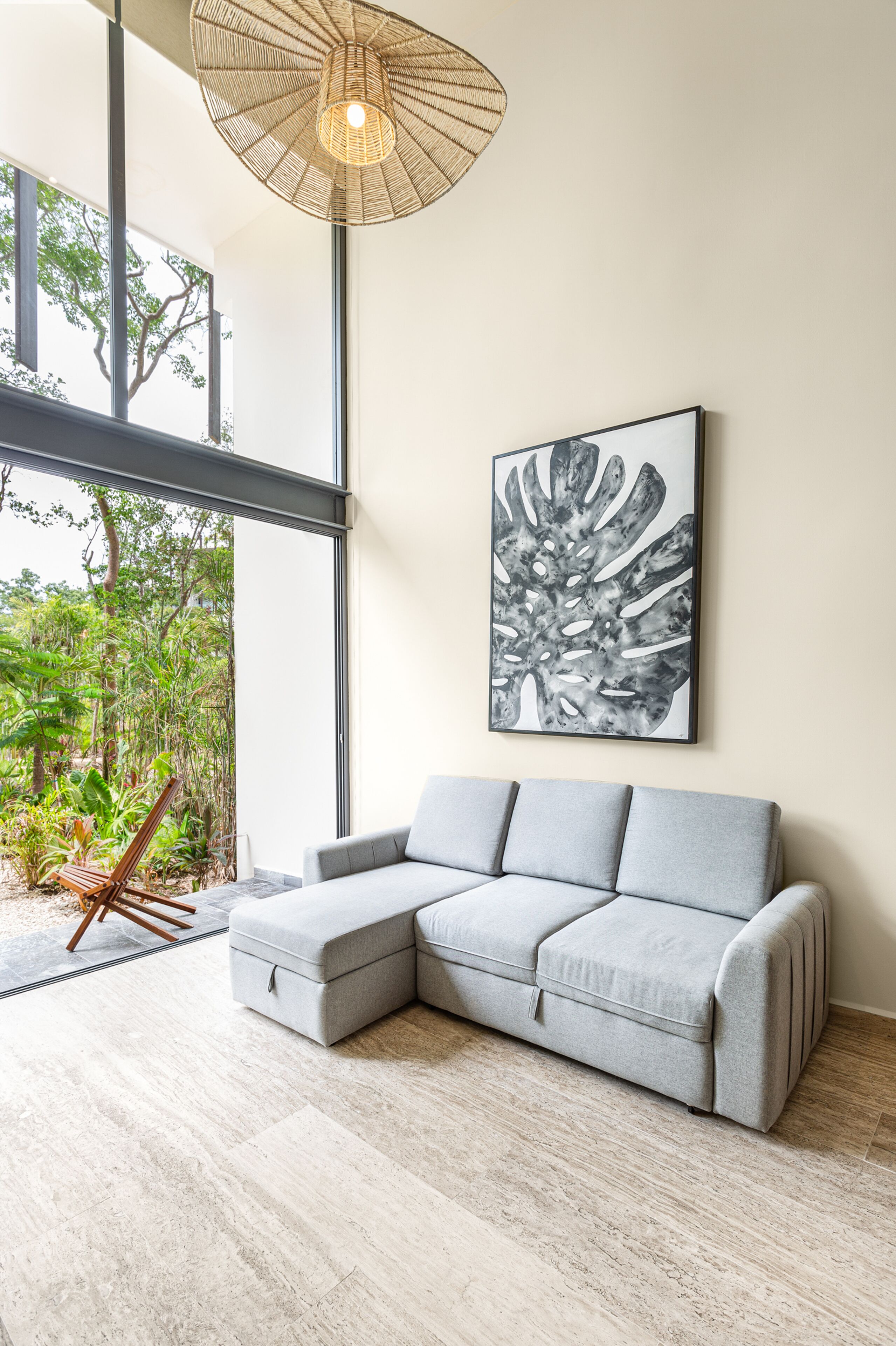 A modern living room with a grey sectional sofa, a large abstract artwork, and a large window offering a view of greenery.