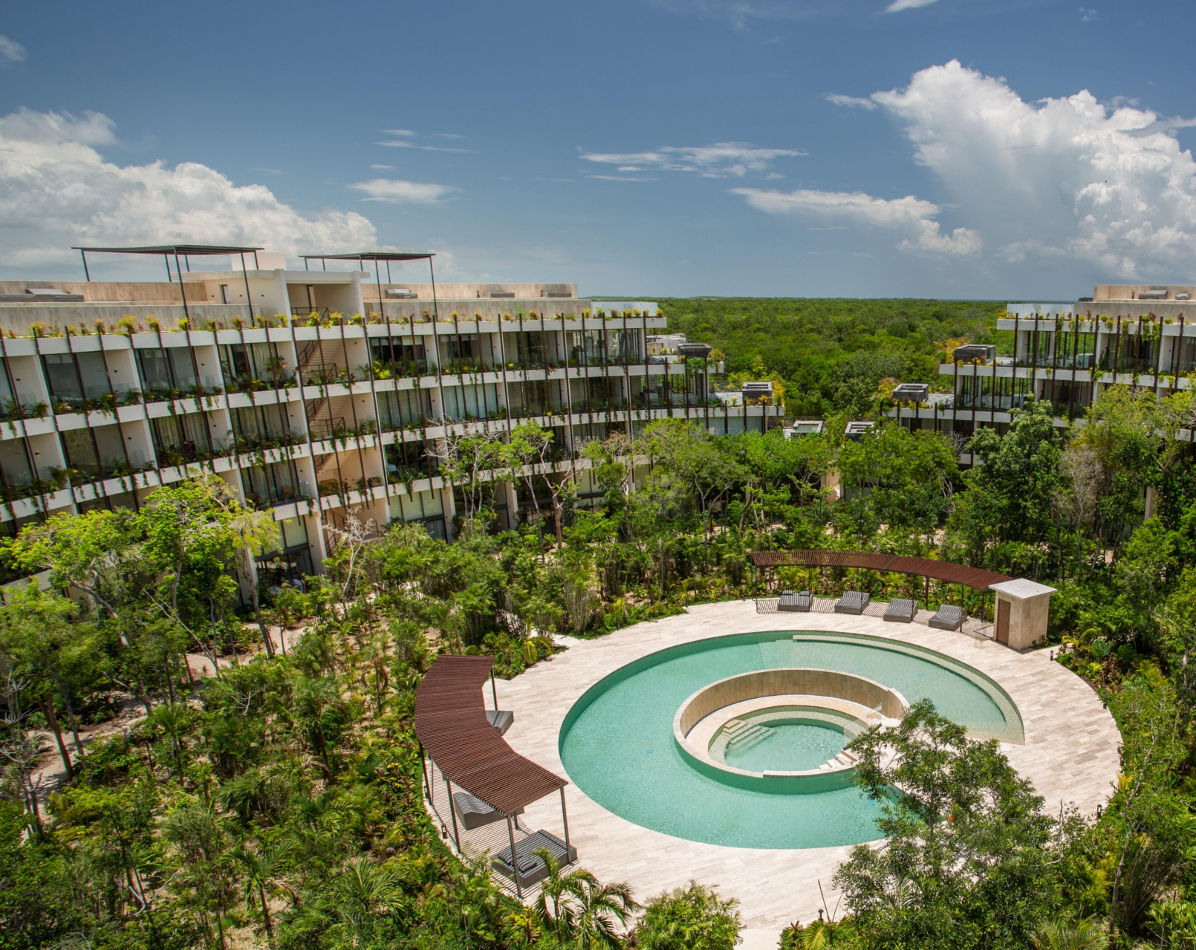 A modern hotel with a circular pool surrounded by lush greenery and a clear blue sky.