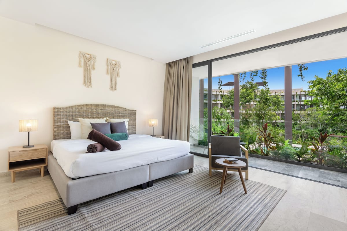Habitación de hotel moderna con una cama grande, mesitas de noche, una silla y vistas a zonas verdes a través de amplios ventanales.