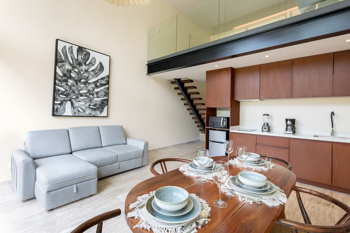 Un espacio habitable moderno con zona de comedor, sofá gris, mesa de comedor de madera y cocina con armarios de madera y una escalera.