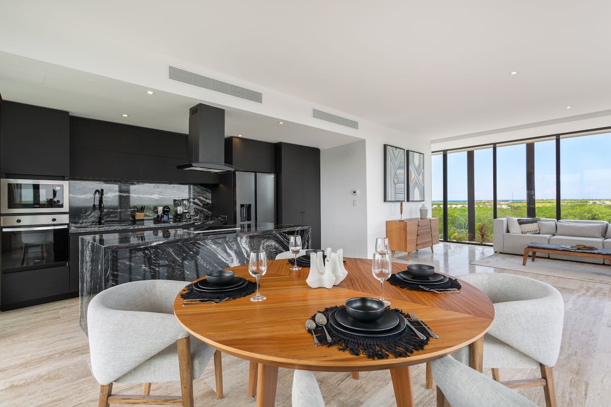 Una cocina moderna con una isla de mármol, electrodomésticos negros y una zona de comedor con mesa de madera y sillas grises.