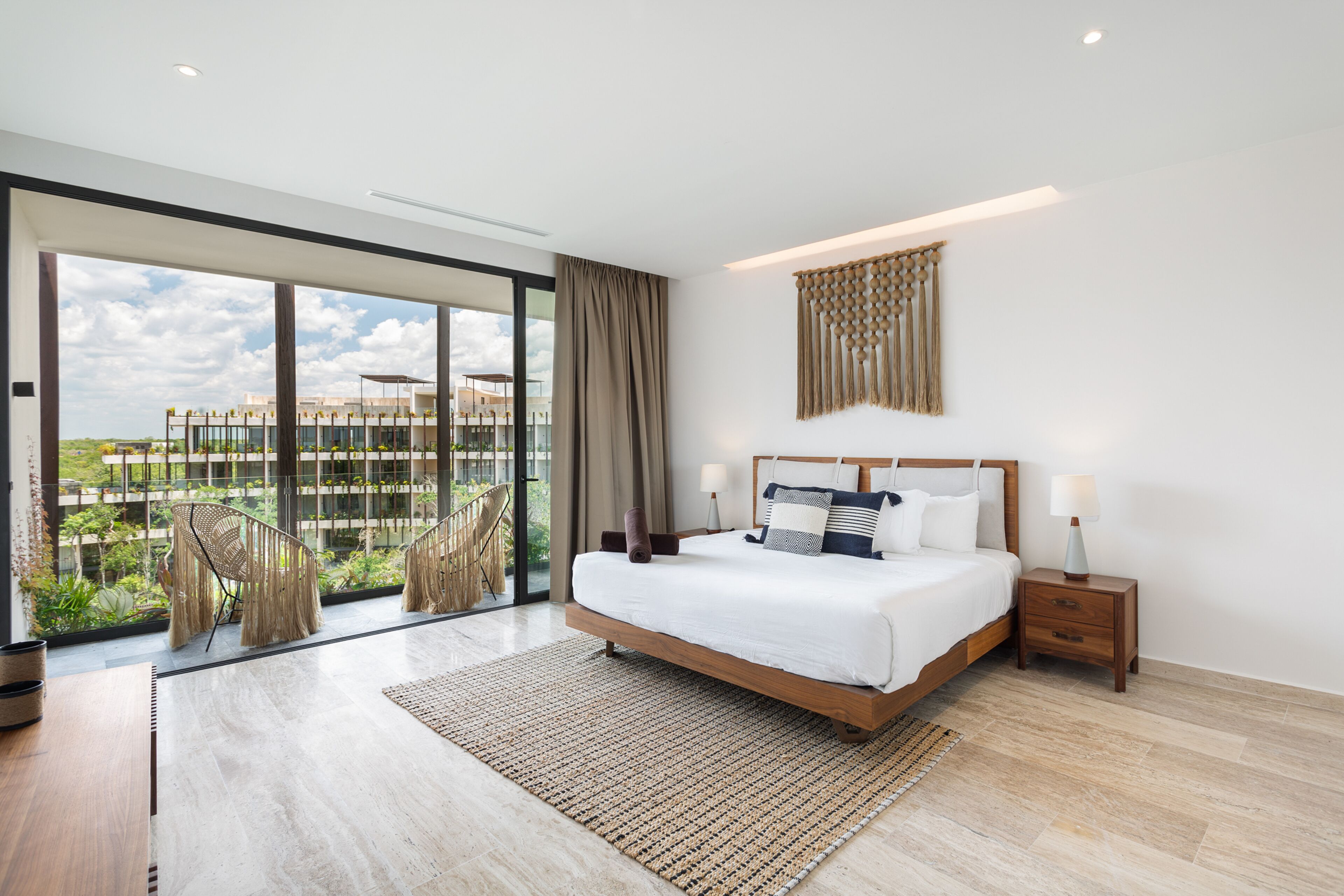 A modern bedroom with a large bed, a wooden nightstand, a bedside table, a large window with a view of buildings, and a woven rug.