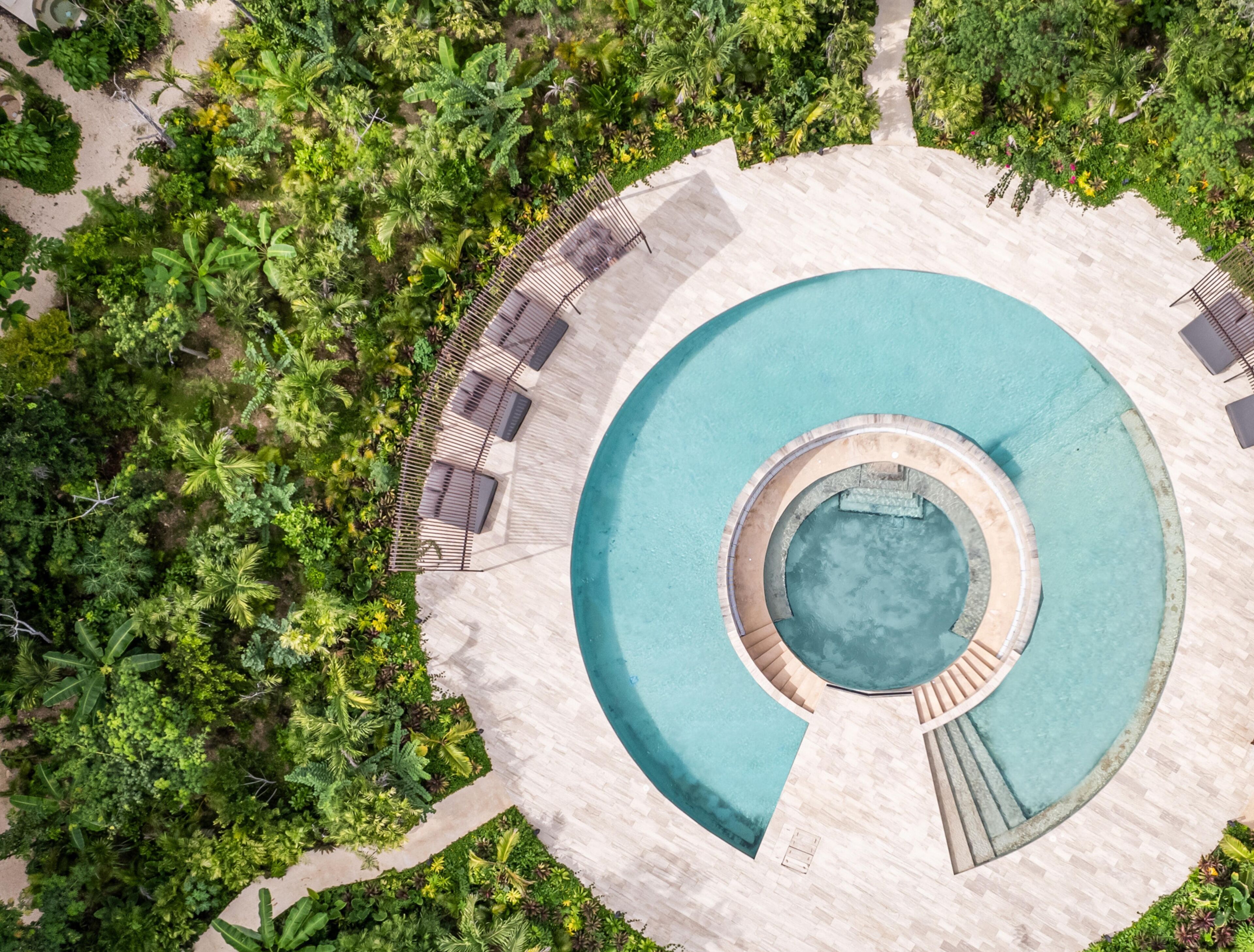 An aerial view of a circular pool surrounded by lush greenery and a sandy area with lounge chairs.
