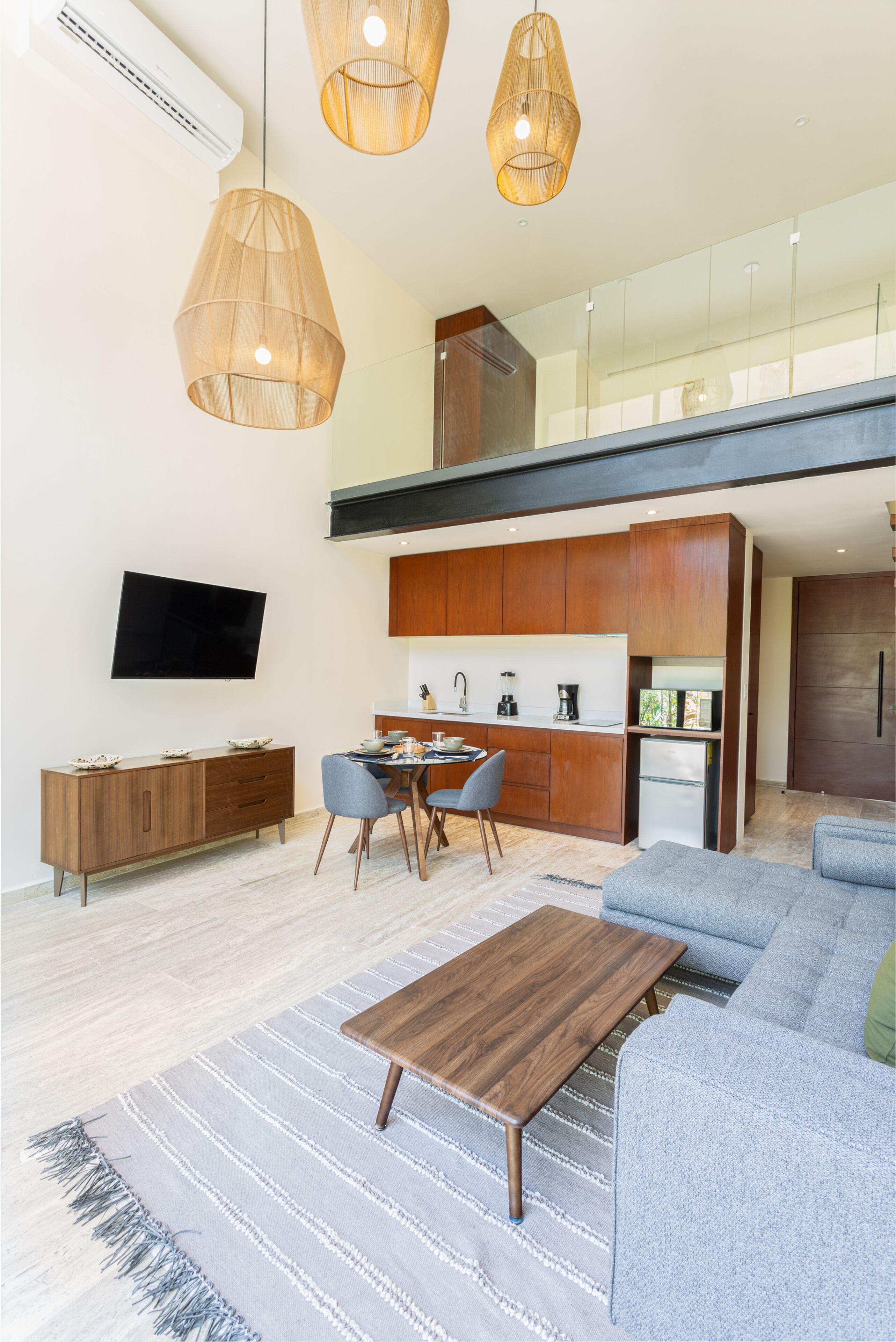 A modern living room with a sofa, a coffee table, and a dining area with chairs. There is a kitchen area with cabinets and a refrigerator. A flat-screen TV is mounted on the wall.