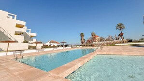 Una piscina con agua cristalina azul, rodeada por un edificio blanco con balcones y palmeras.