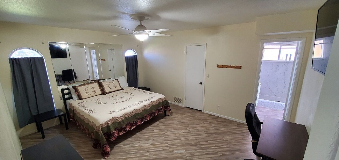 Une chambre à coucher comprenant un lit, un bureau, une chaise, un ventilateur de plafond et une porte donnant sur un balcon.