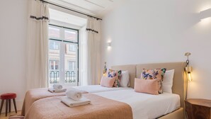 Habitación de hotel con dos camas, un taburete rojo, una mesita de noche de madera y vistas a edificios desde la ventana.