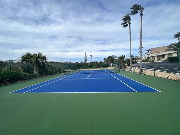 Una cancha de tenis con área verde circundante, palmeras y un edificio al fondo.