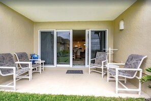 Un patio cubierto con sillas blancas y una mesa, una puerta corrediza de vidrio que da a una habitación y una planta pequeña a la derecha.