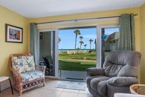 A room with a view of the beach, a wicker chair, a grey sofa, and a small table.