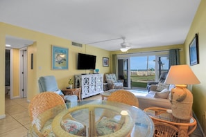 A living room with a glass table, wicker chairs, a sofa, a television, and a view through a sliding glass door.