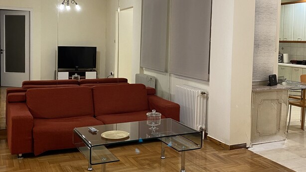 A living room with a sofa, a glass coffee table, a TV, and a kitchen area with a counter and chairs. - An elegant apartment in one of the most central areas of Athens. (Αθήνα)
