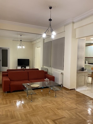 Een woonkamer met een bank, een glazen salontafel, een televisie en een keukenhoek met een aanrecht en stoelen.