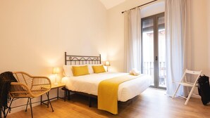Une chambre à coucher avec un lit, deux tables de chevet avec des lampes, un fauteuil en osier, un balcon avec une chaise blanche et un plancher en bois.