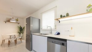 Una cucina moderna con frigorifero in acciaio inossidabile, lavastoviglie e piano di lavoro bianco. C'è un'area pranzo con tavolo e sedie, e una pianta in vaso.