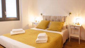 A neatly made bed with a yellow blanket and pillows, two bedside tables with lamps, and a window with blinds. - Bravissimo Sant Martí; Cozy 1-bedroom apartment in Girona's historic center (Girona)
