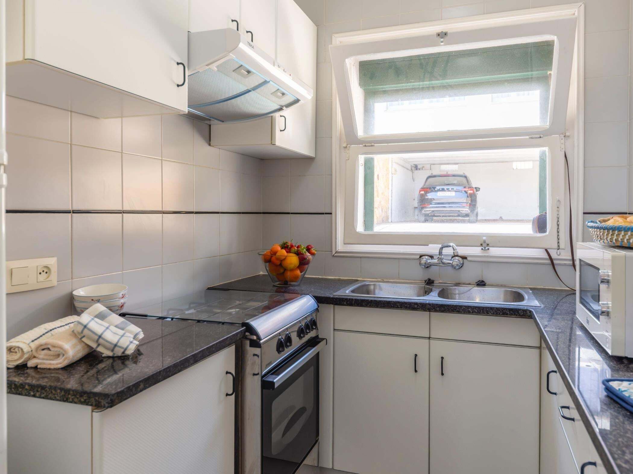 A compact kitchen with a built-in oven, a window with a view of a parked car, and a bowl of fruit on the counter.