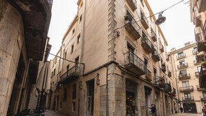 A narrow street with buildings on either side, featuring balconies with plants, a shop entrance, and outdoor seating. - Bravissimo Ferreries, unique 3-bedroom apartment (Girona)