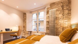 A hotel room with a stone wall, a bed with a striped blanket, a desk with a laptop, and a window with a view of balconies. - Bravissimo Ferreries, unique 3-bedroom apartment (Girona)