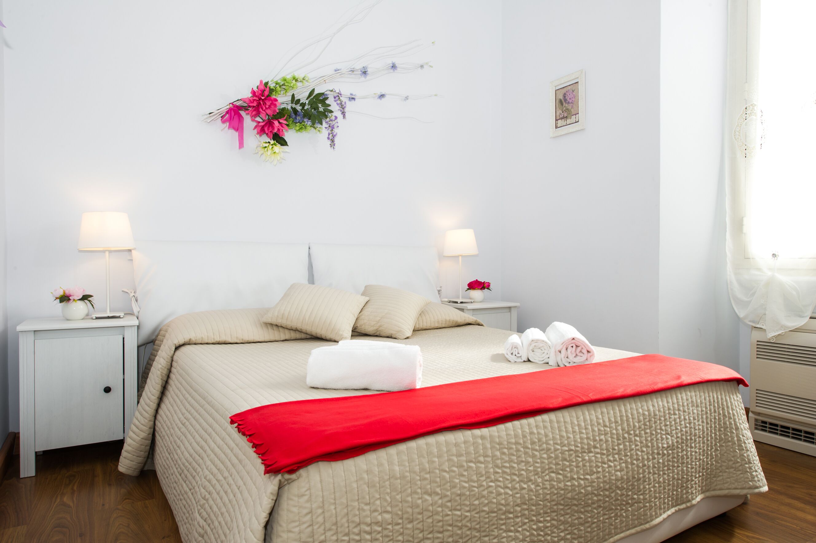 Un lit bien fait, recouvert d’une couverture rouge, deux tables de chevet avec des lampes, une décoration murale à motifs floraux et un petit tableau encadré.