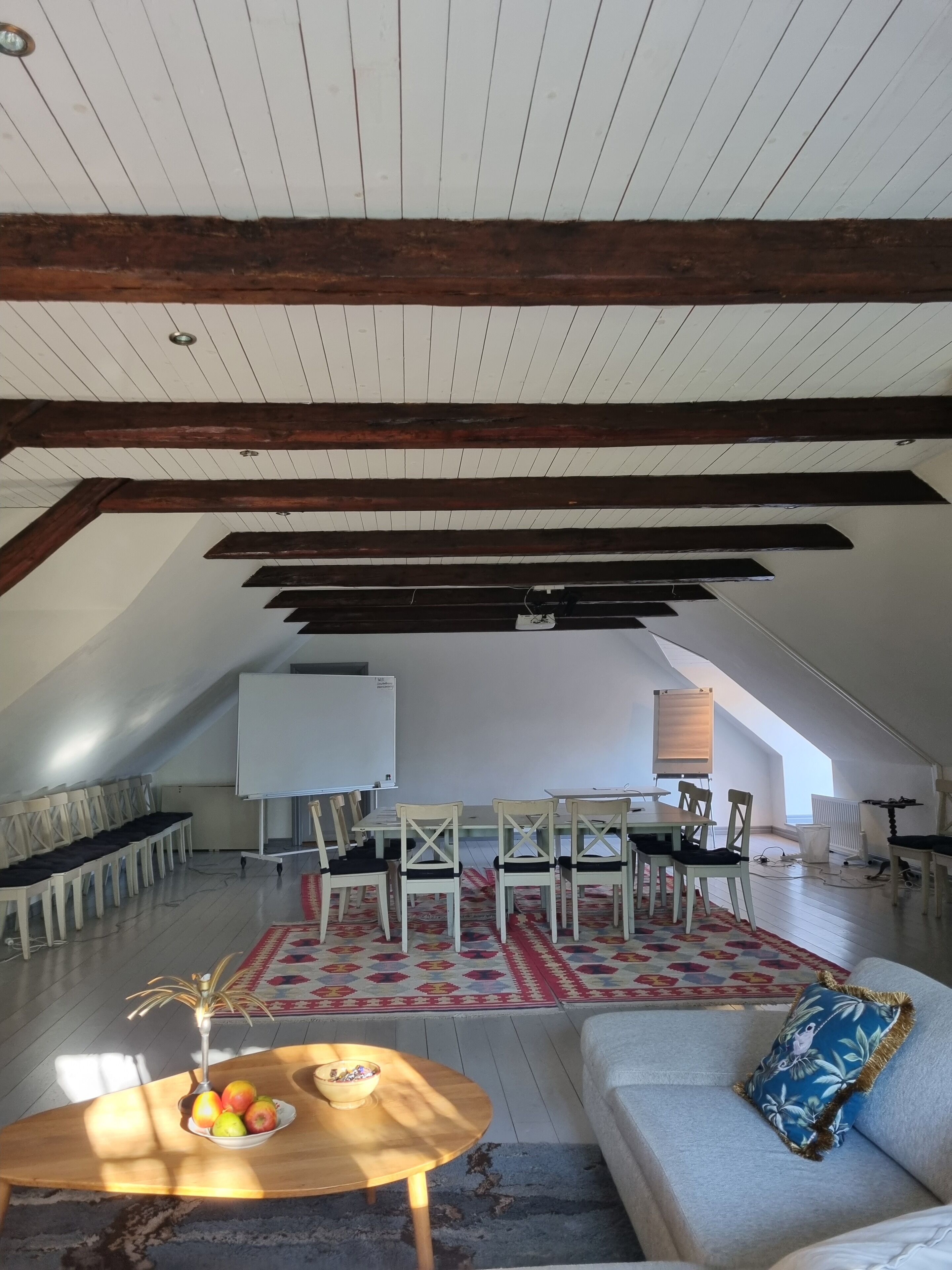 Un amplio ático con techo alto, vigas de madera a la vista, una pizarra, una mesa de comedor con sillas, un sofá y una mesa baja con fruta.