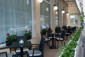 An outdoor cafe with tables and chairs, potted plants, and a view of the street. - Anny Hotel (Jesolo)