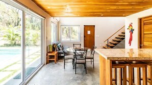 Een modern interieur met een houten plafond, een eethoek met een ronde tafel en stoelen, een keuken met een granieten aanrechtblad en een grote glazen schuindeur die uitkomt bij een zwembad.