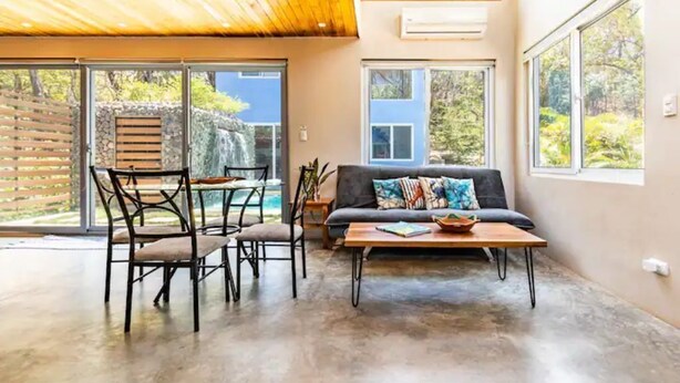 Un vaste espace de vie doté d’un plafond en bois, de grandes fenêtres, d’un canapé, d’une table basse et d’une table à manger avec des chaises.