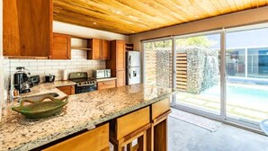 Une cuisine moderne avec des meubles en bois, des plans de travail en granit et une grande porte-fenêtre coulissante donnant sur une zone aménagée autour d’une piscine.