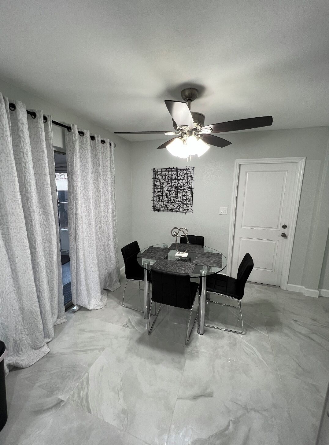 A dining area with a glass table and four chairs, a ceiling fan, and a door in the background.