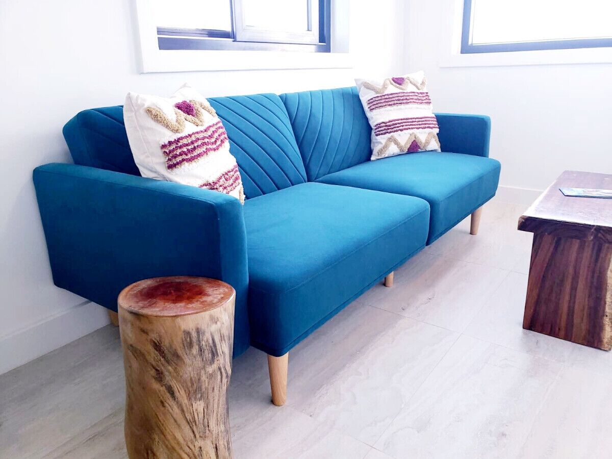 A blue sofa with patterned cushions, a wooden stool, and a small wooden table in a bright room with white walls and a window.