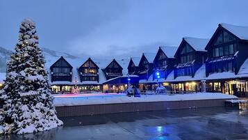 A snow-covered outdoor area with a Christmas tree, a building with multiple windows and a ski and snowboard rental sign, and a wooden deck.