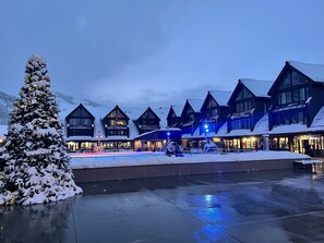 Un'area esterna innevata con un albero di Natale, un edificio con diverse finestre e un cartello che indica il noleggio di sci e snowboard, e un molo in legno.