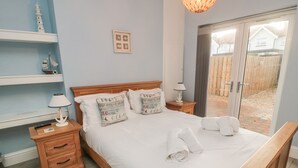 A bedroom with a wooden bed, two bedside tables, a nightstand, a lamp, a shelf with decorative items, and a view of the outside through a glass door. - Bamburgh Dunes (Bamburgh)