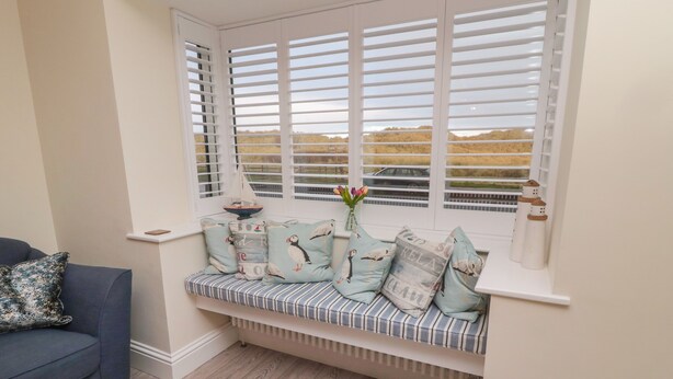 A cozy seating area with a cushioned bench, a blue sofa, and a window with white shutters. - Bamburgh Dunes (Bamburgh)