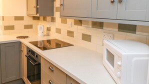 A modern kitchen with grey cabinets, a black induction cooktop, a white microwave, and a small oven. - Firkin Cottage (Bedale)