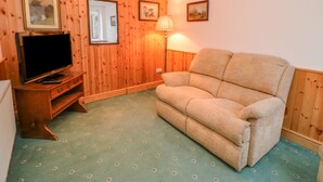 A room with wooden panel walls, a flat-screen TV on a wooden stand, a beige sofa, a floor lamp, and a patterned carpet. - Firkin Cottage (Bedale)