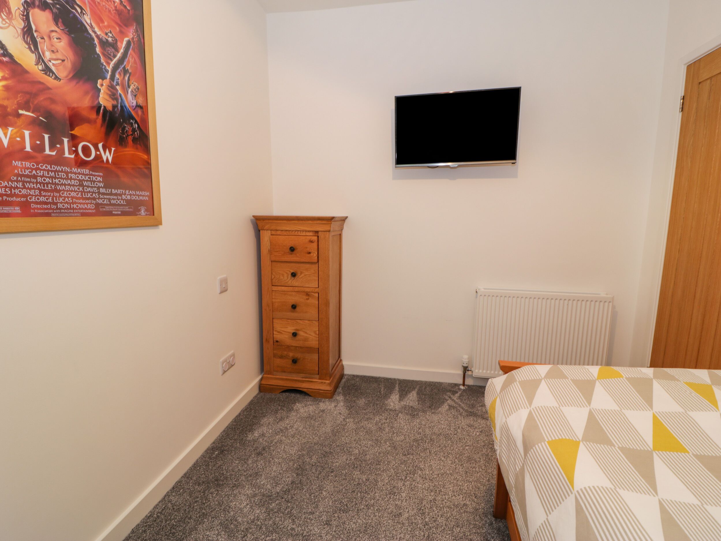 A bedroom with a bed, a wooden dresser, a television on the wall, and a poster on the wall.