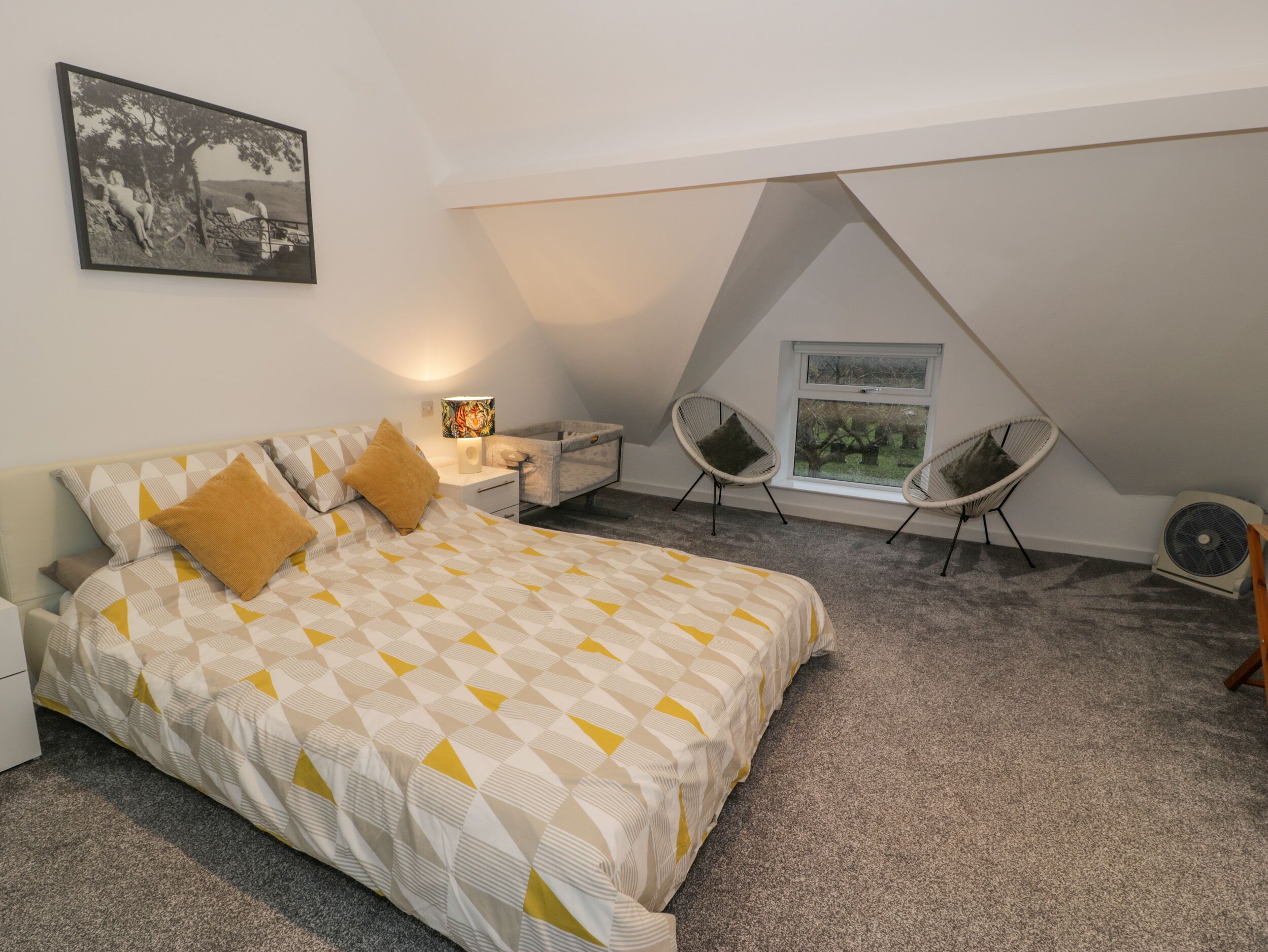 A bedroom with a bed, nightstands, a lamp, a crib, two chairs, a window, and a fan.