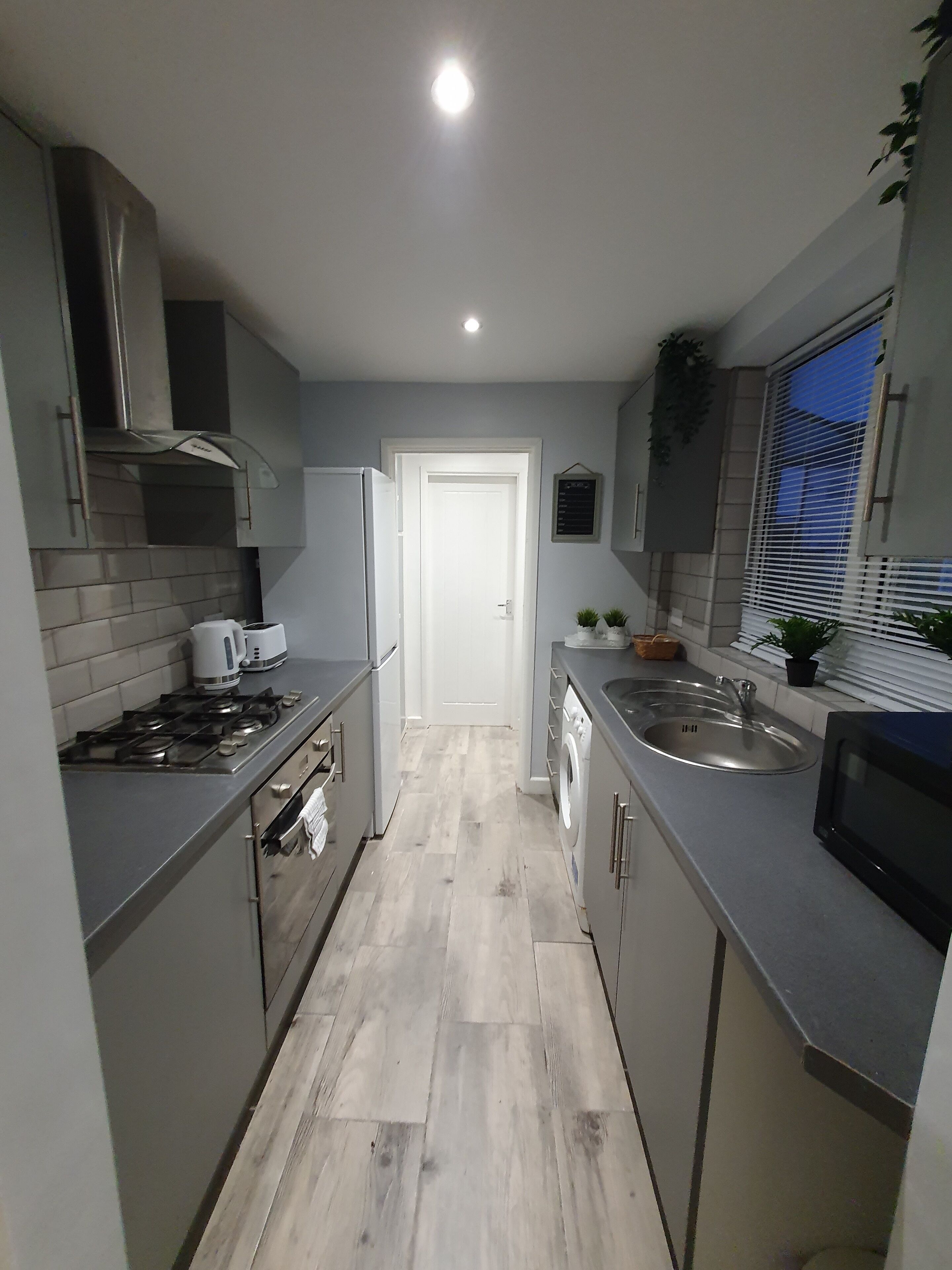 A narrow kitchen with grey cabinets, a stainless steel sink, a microwave, and a stove.