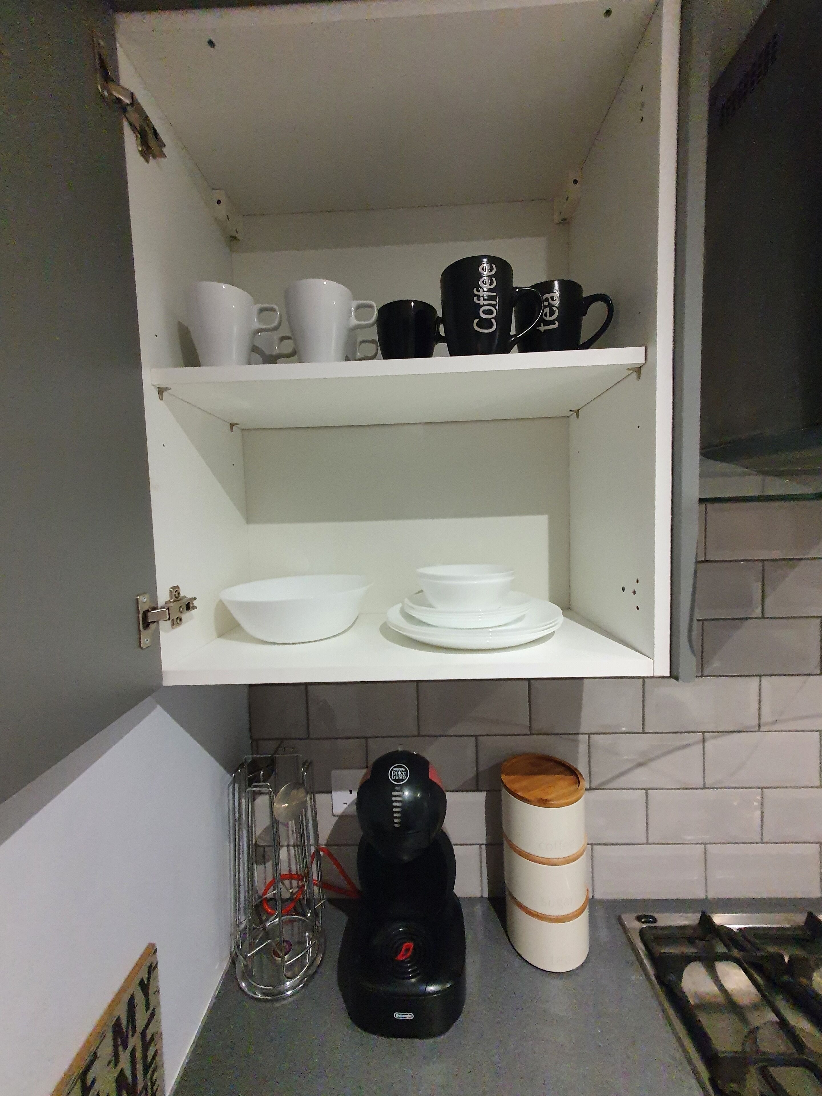 A kitchen with a white cabinet containing white and black mugs, a coffee maker, and a kettle.