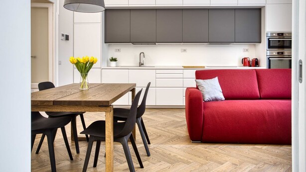 Une cuisine moderne avec des meubles blancs, une table à manger en bois, des chaises noires, un canapé rouge et un luminaire suspendu.