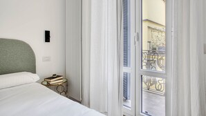 Une chambre à coucher avec un lit, une table de chevet avec des livres, un radiateur et une vue sur un balcon doté d’une balustrade.