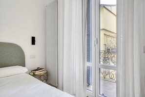 Un dormitorio con cama, una mesita de noche con libros, un radiador y vistas a un balcón con barandilla.