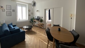 A modern living room with a blue sofa, a wooden dining table with chairs, a TV on a stand, and a large window. - Appart Privé Deluxe Strasbourg City (Strasbourg)